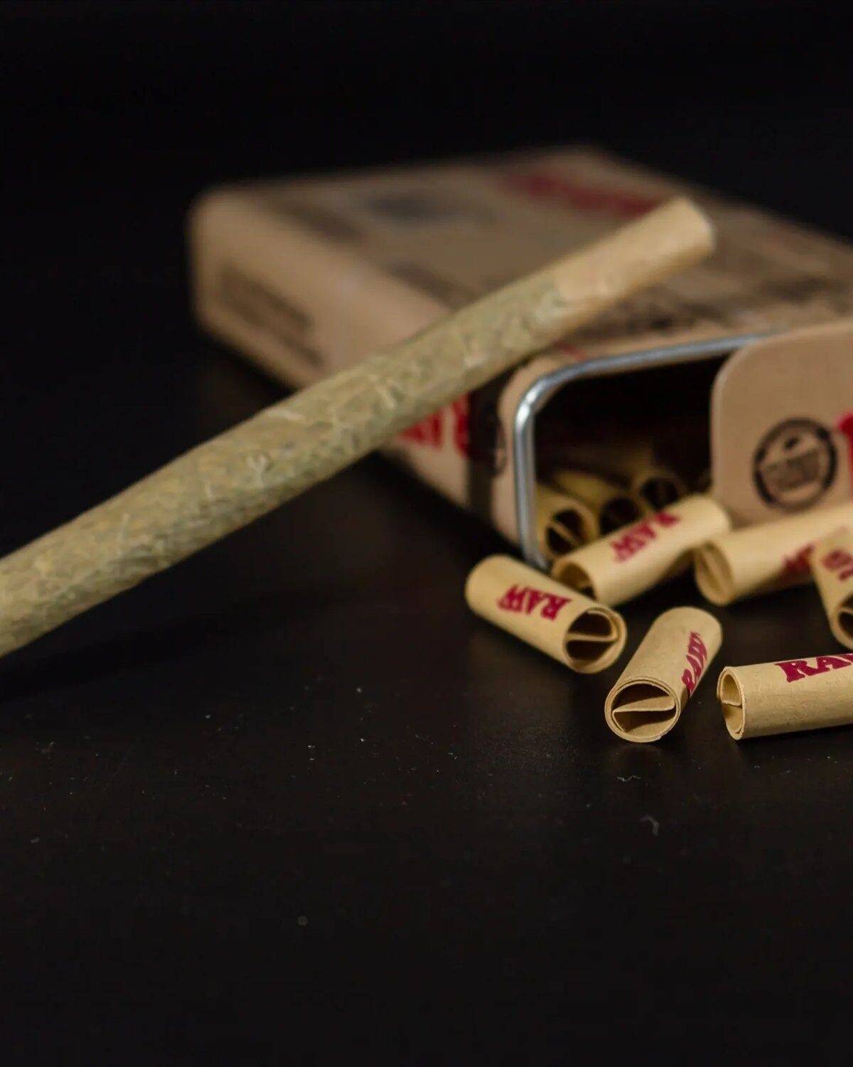 A rolled joint resting against an open tin of RAW rolling papers and pre-rolled tips.